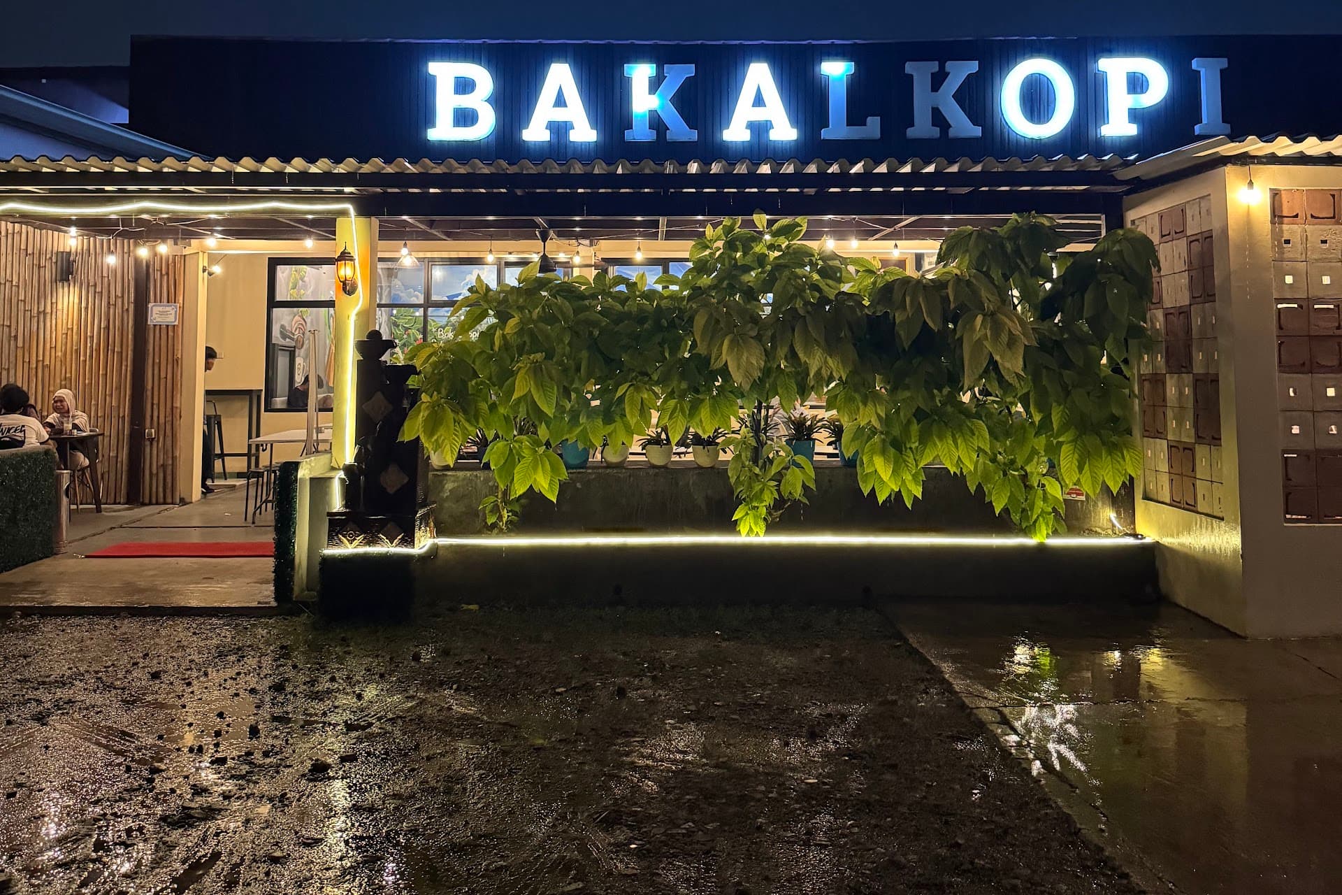 Fasad BAKALKOPI pada malam hari: papan nama menyala, tanaman tropis, dan lampu gantung hangat