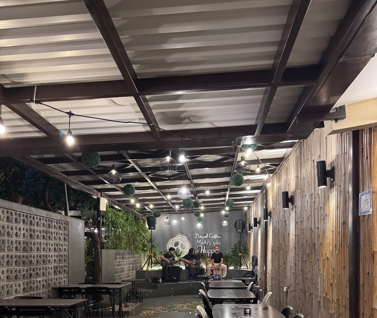 Musisi tampil akustik di teras BAKALKOPI dengan lampu untaian dan dinding bambu